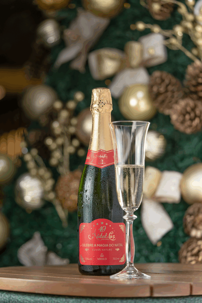 Garrafa do espumante Miolo Cuvée Nature Edição Especial Natal Luz 40 Anos com taça ao lado, em frente a uma árvore de Natal decorada com bolas douradas e pinhas.