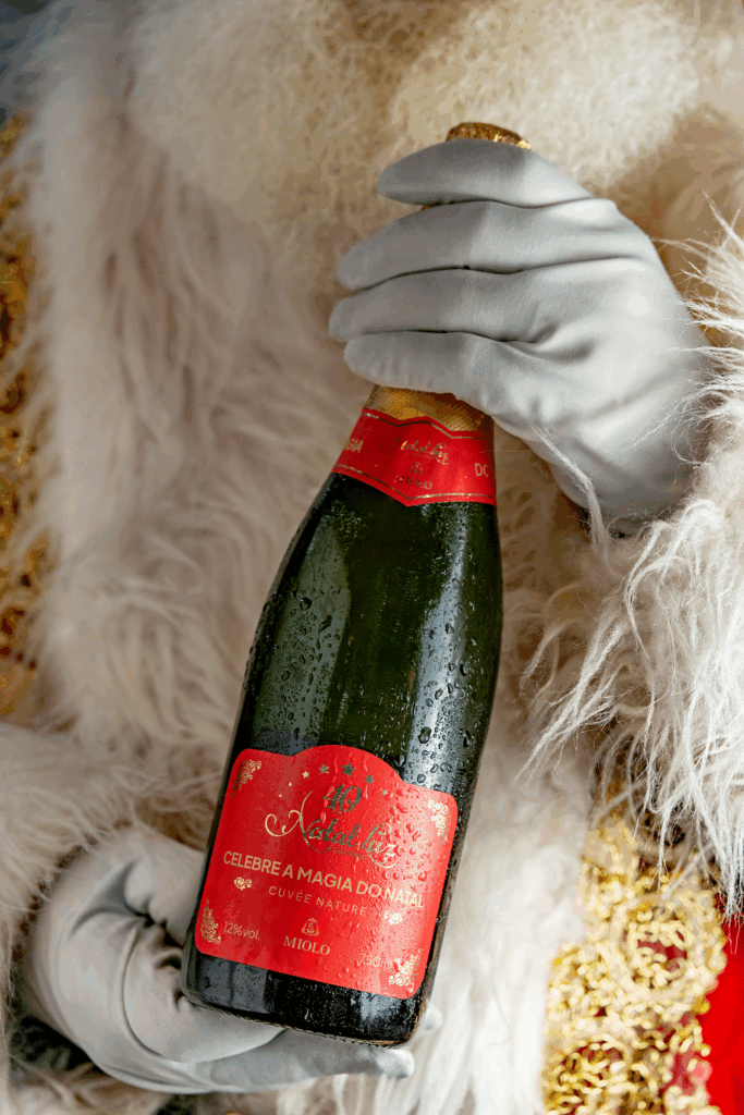 Papai Noel segurando a garrafa do espumante Miolo Cuvée Nature Edição Especial Natal Luz 40 Anos, com detalhes dourados e fundo festivo. Crédito da imagem: divulgação Miolo.
