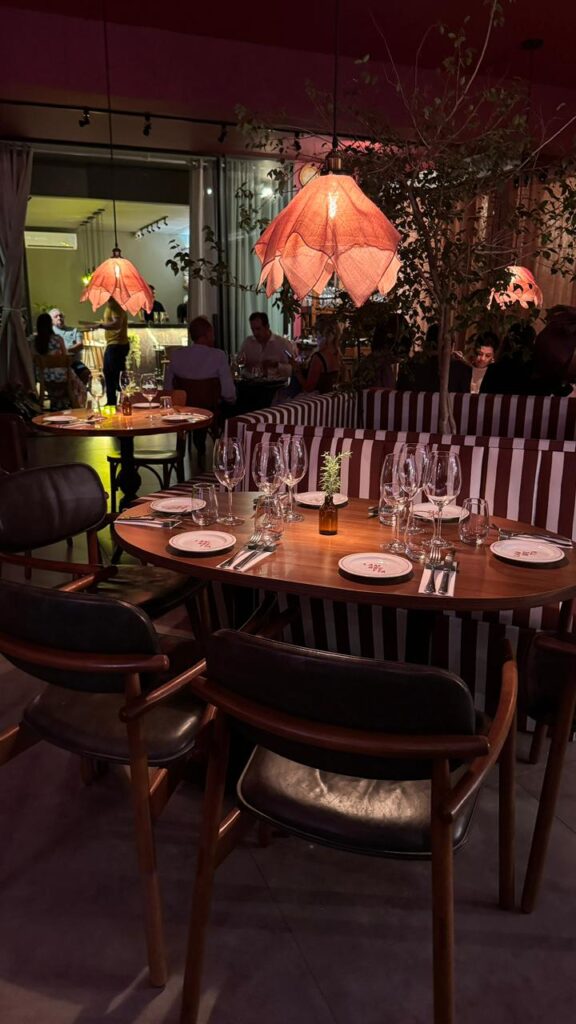 Mesa posta com utensílios de restaurante, copos e taças, claros, em ambiente com iluminação baixa.