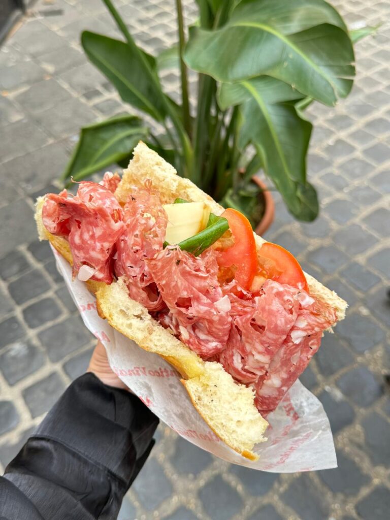 Close do sanduíche All’Antico Vinaio com salame, queijo e tomate sobre pão toscano, sendo segurado por uma mão.
