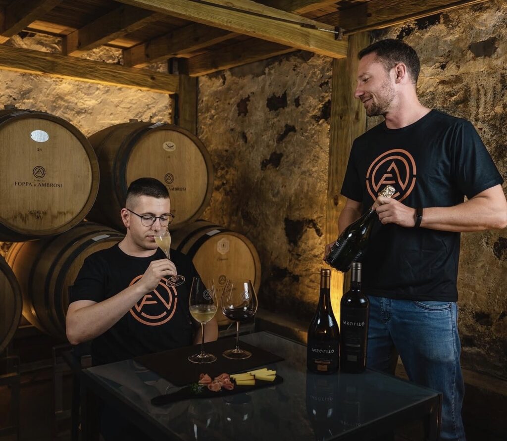 Rodrigo Foppa e Lucas Ambrosi em uma cave no Napa Valley, um servindo vinho enquanto o outro degusta diante de barricas, com garrafas e queijos sobre a mesa.
