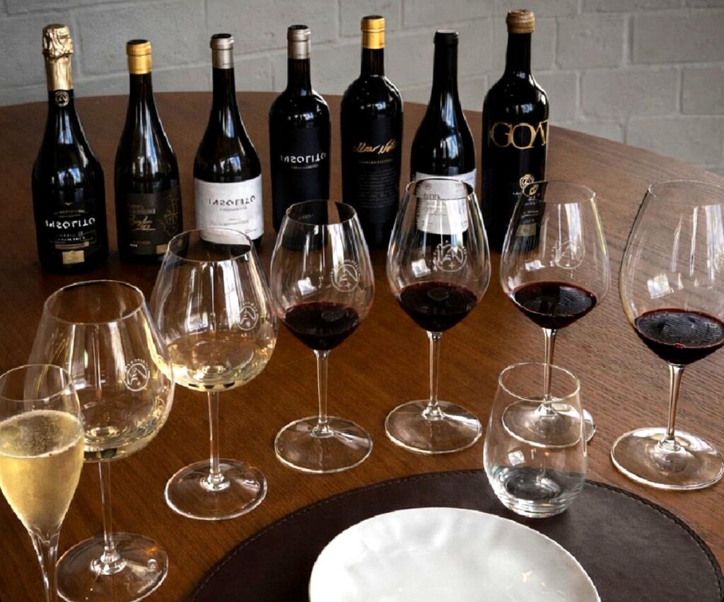 Taças de vinho dispostas sobre mesa de madeira durante degustação da Foppa & Ambrosi, com diferentes estilos de vinhos servidos e garrafas ao fundo, incluindo rótulos como Insólito e GOAT.